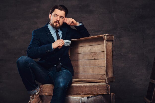 Homme d'affaires pensif d'âge moyen avec des cheveux et une barbe élégants vêtus de jeans, veste et cravate, assis sur des caisses en bois et tenant une tasse de café dans un studio. Isolé sur un fond texturé sombre.
