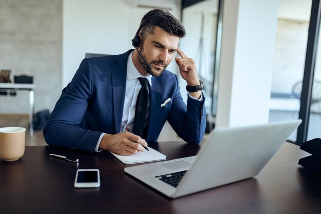 Homme d'affaires pensif prenant des notes tout en travaillant sur un ordinateur portable au bureau