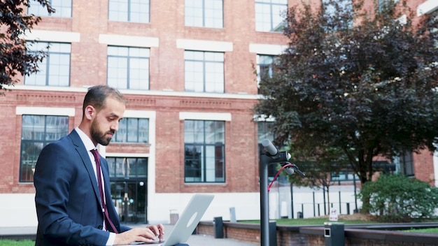 Photo gratuite homme d'affaires prospère en costume travaillant à une présentation marketing assis sur un banc à l'extérieur devant le bureau du bâtiment de l'entreprise. directeur exécutif à la recherche d'informations sur internet.