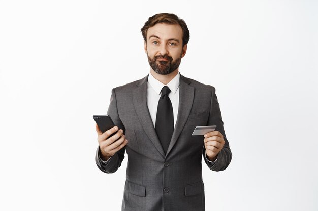 Homme d'affaires sceptique en costume grimaçant déçu tenant une carte de crédit et un téléphone portable debout sur fond blanc