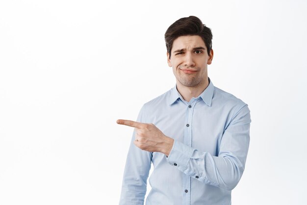 Homme d'affaires sceptique déçu sourire narquois et froncer les sourcils contrarié pointant le doigt vers la gauche à quelque chose de mauvais regard douteux debout en chemise sur fond blanc