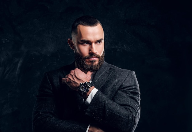 Un Homme D'affaires Sceptique Sérieux Avec Une Barbe Et Des Vêtements Sombres Se Tient Dans Un Studio Photo Sombre.