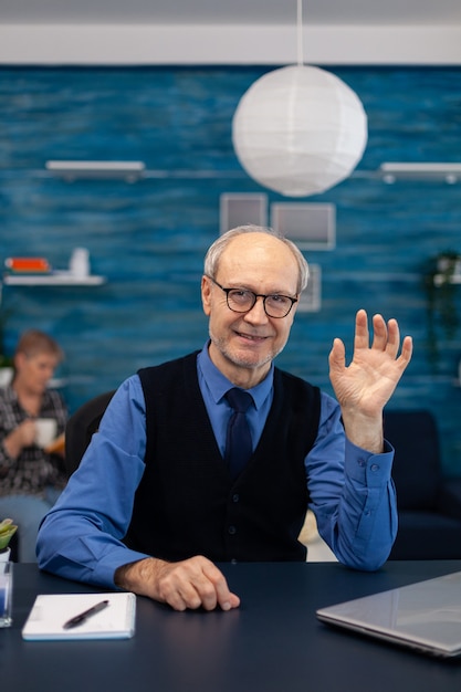 Homme d'affaires senior intelligent travaillant sur ordinateur portable portant une cravate et des lunettes. Homme âgé entrepreneur sur son lieu de travail à domicile utilisant un ordinateur portable assis au bureau pendant que sa femme tient la télécommande de la télévision.