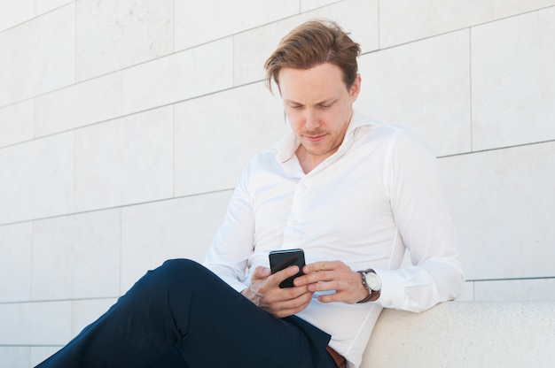 Homme d'affaires sérieux à l'aide de smartphone sur un banc à l'extérieur