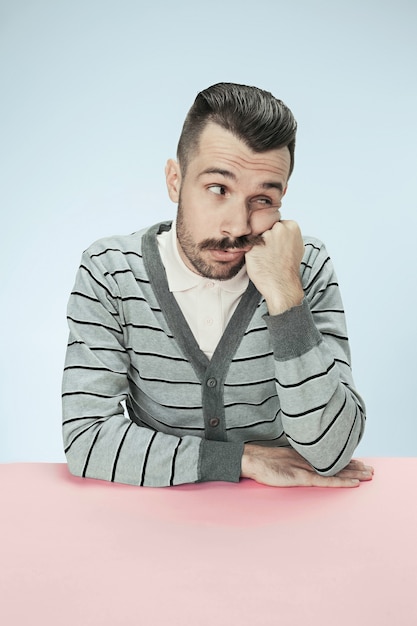 Homme d'affaires sérieux, ennuyé et terne assis à table sur fond bleu studio. Le portrait dans un style minimalisme