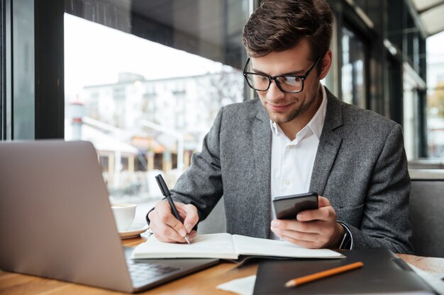 Homme d'affaires souriant à lunettes assis près de la table au café avec un ordinateur portable tout en utilisant un smartphone et en écrivant quelque chose