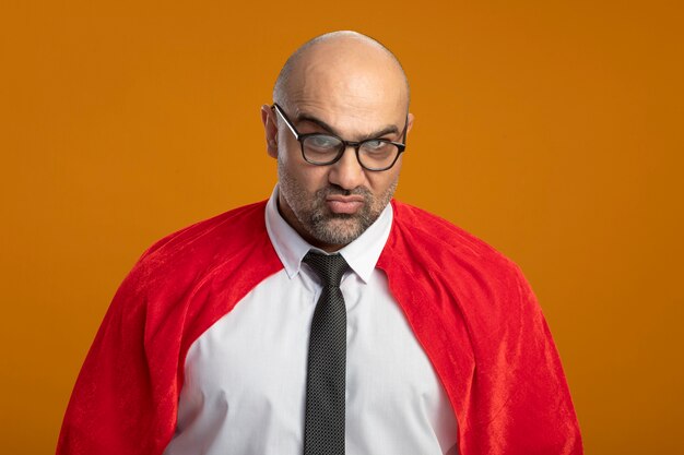 Homme d'affaires de super héros en cape rouge et lunettes à l'avant d'être mécontent de froncer les sourcils debout sur un mur orange