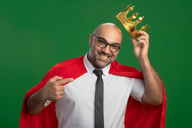 Homme d'affaires de super héros en cape rouge et lunettes portant couronne à la recherche de pointign souriant confiant avec index à la couronne debout sur le mur vert