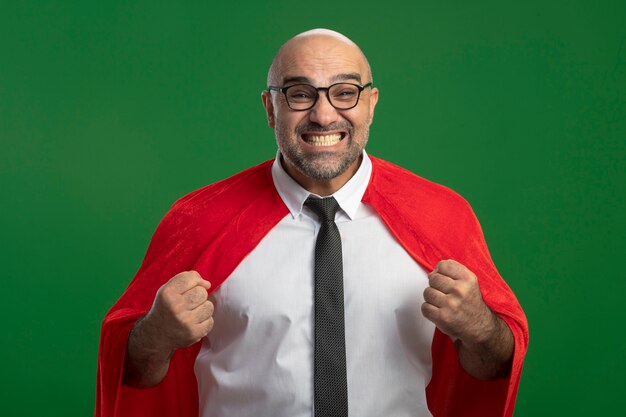 Homme d'affaires de super héros en cape rouge et lunettes serrant le poing heureux et excité debout sur le mur vert