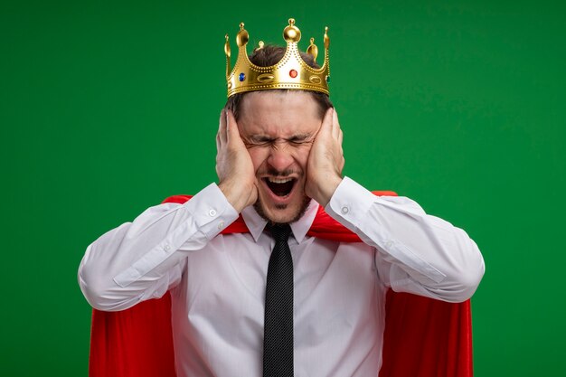 Photo gratuite homme d'affaires de super héros en cape rouge portant la couronne en criant d'être frustré avec les mains sur son visage avec les yeux fermés debout sur le mur vert