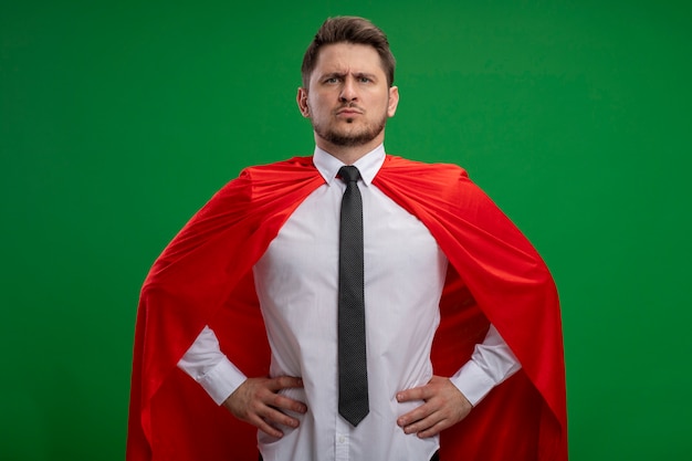 Homme d'affaires de super héros en cape rouge regardant la caméra avec une expression confiante sérieuse avec les bras à la hanche debout sur fond vert