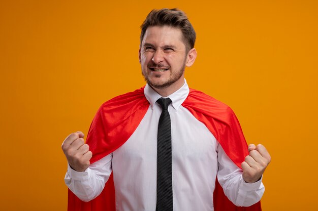 Homme d'affaires de super héros en cape rouge serrant les poings heureux et excité debout sur le mur orange