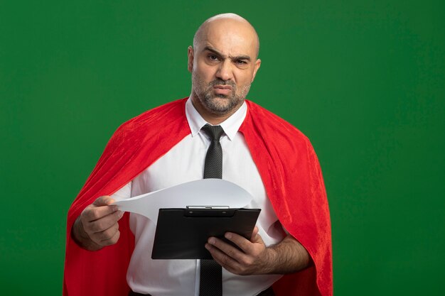 Homme d'affaires de super héros en cape rouge tenant le presse-papiers avec des pages blanches à l'avant avec un visage sérieux fronçant les sourcils debout sur un mur vert