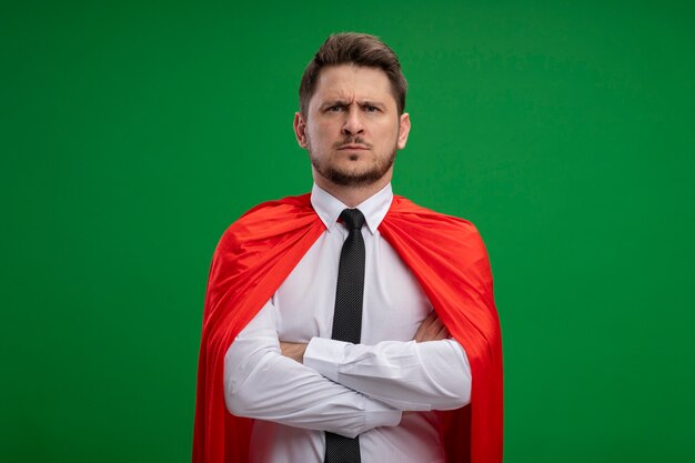 Homme d'affaires de super héros en cape rouge avec un visage sérieux avec les bras croisés sur la poitrine debout sur le mur vert