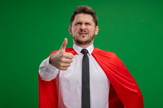 Homme d'affaires de super héros en cape rouge avec visage sérieux montrant les pouces vers le haut debout sur le mur vert