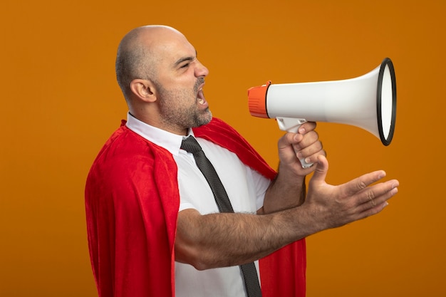 Homme d'affaires de super-héros en colère en cape rouge criant au mégaphone avec bras debout sur un mur orange