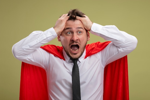 Homme d'affaires de super héros fou fou en cape rouge criant avec une expression agressive tirant ses cheveux se déchaînant debout sur le mur vert