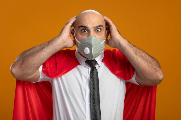 Photo gratuite homme d'affaires de super-héros fou en masque facial protecteur et cape rouge avec un regard étonné fou de surprise tenant les mains sur sa tête