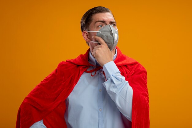 Homme d'affaires de super héros en masque facial protecteur et cape rouge à côté avec une expression pensive sur le visage avec la main sur le menton pensant debout sur fond orange