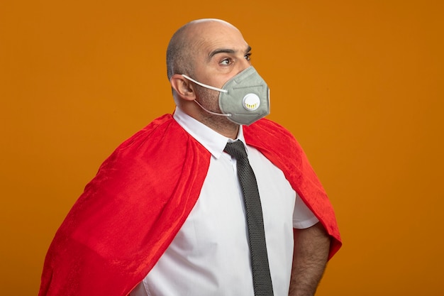 Homme d'affaires de super héros en masque facial de protection et cape rouge à côté avec une expression confiante sérieuse debout sur un mur orange