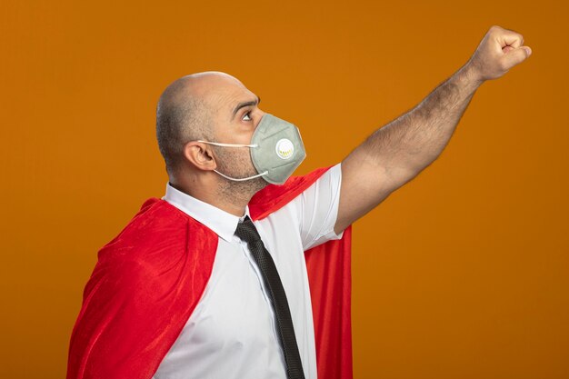 Homme d'affaires de super héros en masque facial de protection et cape rouge faisant le geste gagnant à la confiance debout sur le mur orange