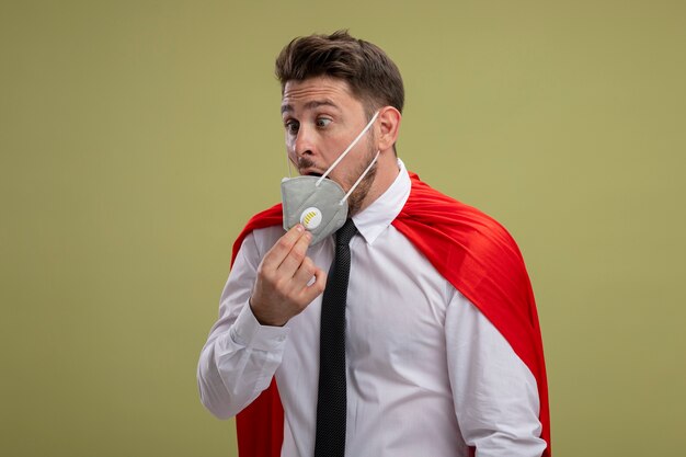 Homme d'affaires de super héros en masque facial de protection et cape rouge à la prise de masque confus debout sur fond vert