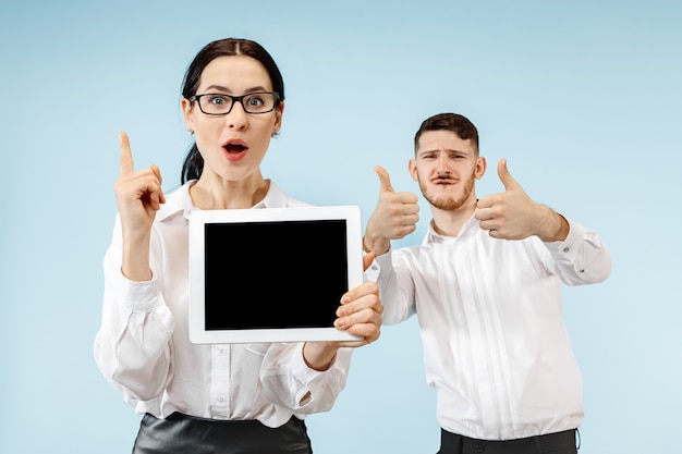 L'homme d'affaires surpris et femme souriant sur un fond de studio bleu et montrant un écran vide d'ordinateur portable ou de tablette
