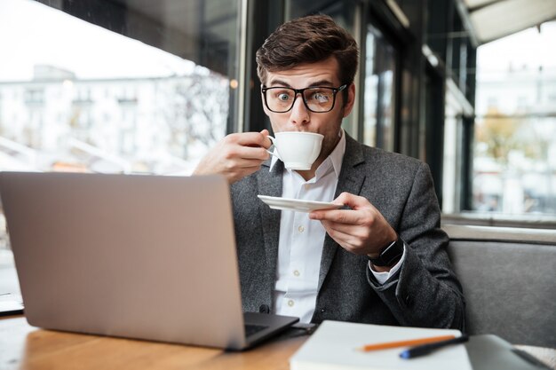 Homme d'affaires surpris à lunettes assis près de la table au café avec ordinateur portable tout en buvant du café et en regardant