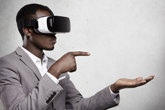 Photo gratuite homme africain attrayant en tenue de soirée et lunettes 3d pointant ses doigts sur l'espace de copie au-dessus de sa paume ouverte comme s'il utilisait un gadget.