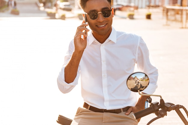 Homme africain sans soucis à lunettes de soleil assis sur une moto moderne à l'extérieur et parler par le smartphone