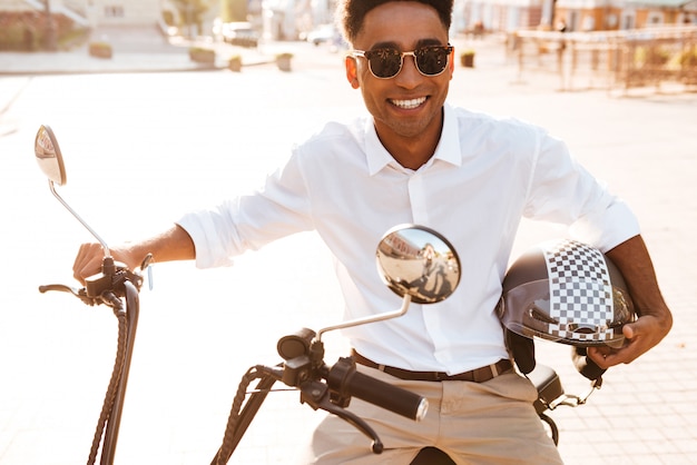 Homme africain souriant assis sur une moto moderne à l'extérieur et en regardant la caméra