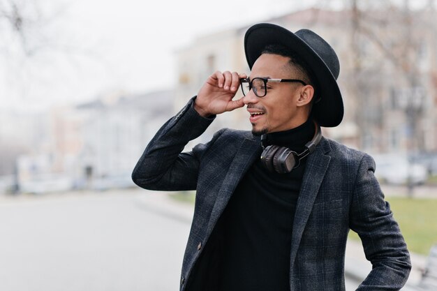 Homme africain souriant en veste de laine touchant ses lunettes et regardant autour de lui. Heureux jeune homme à la peau brune profitant du beau temps en plein air.