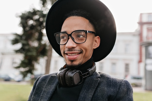 Homme africain spectaculaire avec un sourire sincère posant. Photo extérieure d'un modèle masculin noir mignon dans des lunettes et des écouteurs.