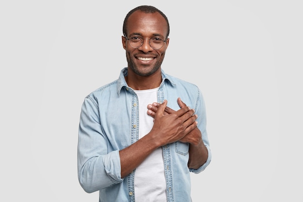 Homme africain touché et reconnaissant tient les mains sur la poitrine, exprime sa gratitude, regarde agréablement la caméra