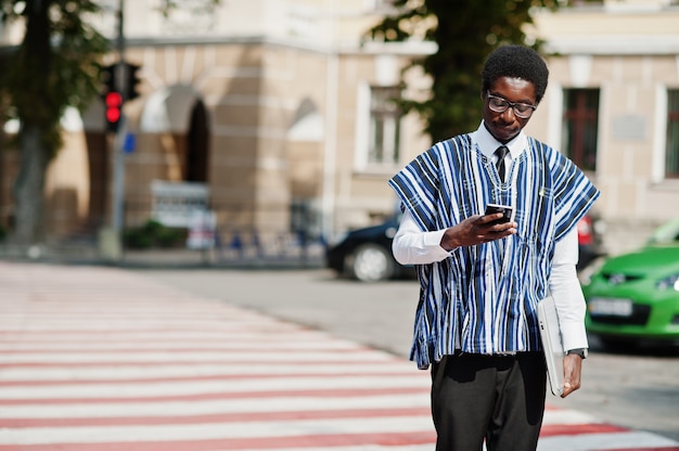 Homme africain en vêtements traditionnels et lunettes marchant au passage pour piétons avec téléphone portable et ordinateur portable afrique affaires.