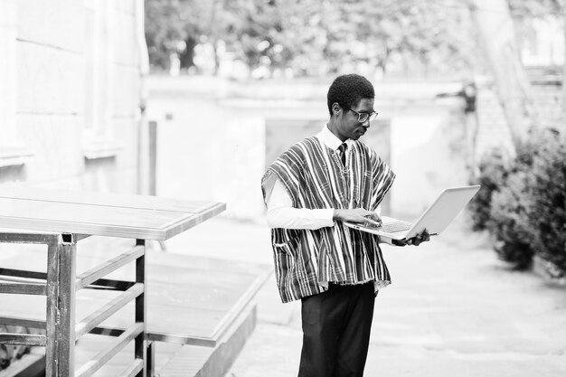 Homme africain en vêtements traditionnels et lunettes avec ordinateur portable travaillant en plein air