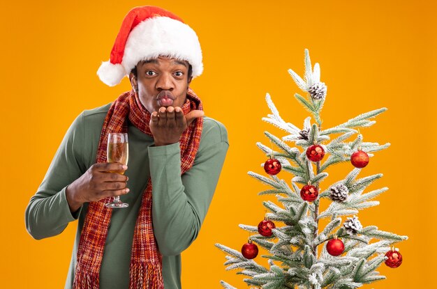 Homme afro-américain en bonnet de noel et écharpe autour du cou tenant une coupe de champagne soufflant un baiser avec le bras devant son visage debout à côté d'un arbre de noël sur fond orange