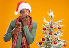 Photo gratuite homme afro-américain en bonnet de noel et écharpe autour du cou tenant un verre de champagne regardant la caméra choqué couvrant la bouche avec la main à côté de l'arbre de noël sur fond orange