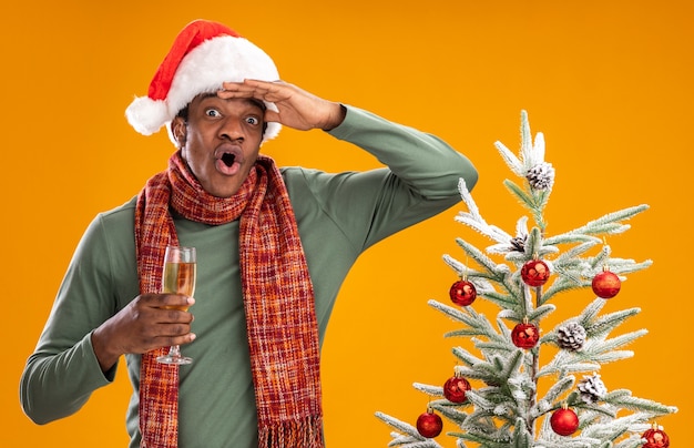 Photo gratuite homme afro-américain en bonnet de noel et écharpe autour du cou tenant un verre de champagne à la surprise de loin avec la main sur la tête debout à côté d'un arbre de noël sur fond orange