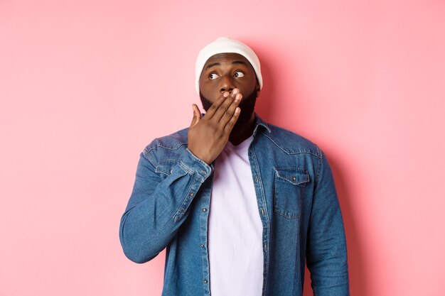 Un homme afro-américain choqué haletant, regardant à gauche avec admiration, bavardant, debout sur fond rose