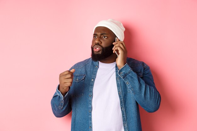 Un homme afro-américain en colère parle au téléphone et menace quelqu'un avec le poing levé, l'air dérangé par la personne, debout sur fond rose.