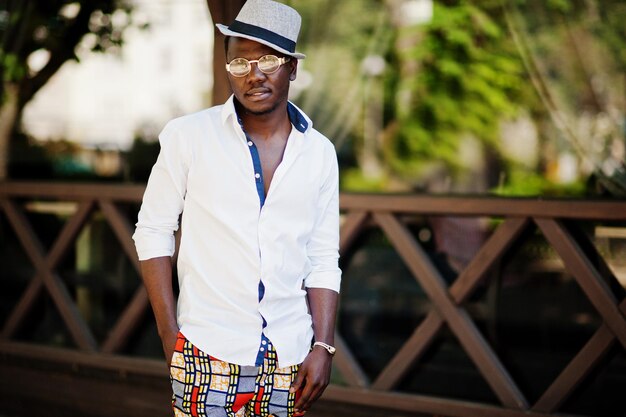 Homme afro-américain élégant en chemise blanche et pantalon coloré avec chapeau et lunettes posés en plein air garçon modèle à la mode noir