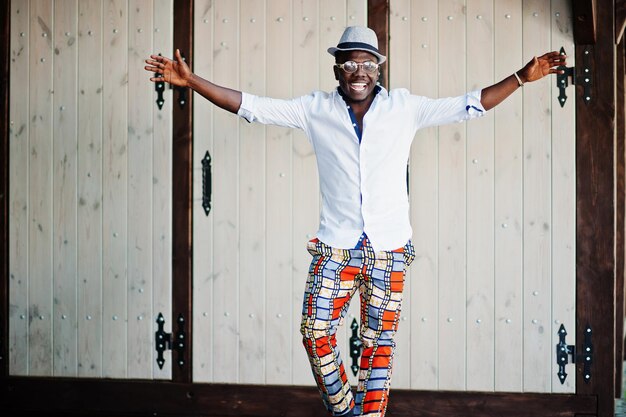 Homme afro-américain élégant en chemise blanche et pantalon coloré avec chapeau et lunettes posés en plein air garçon modèle à la mode noir