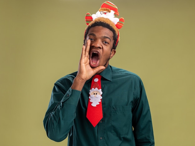 Homme afro-américain émotionnel avec drôle de santa jante et cravate rouge regardant la caméra en criant avec la main près de la bouche debout sur fond vert