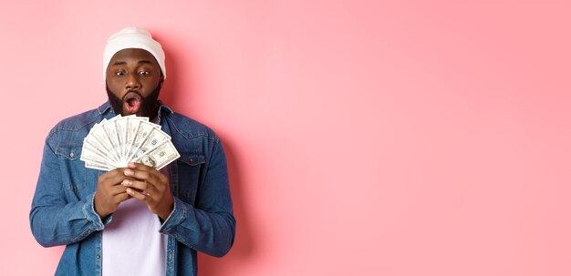 Photo gratuite un homme afro-américain étonné reçoit un prix en espèces montrant de l'argent et regardant avec admiration debout sur ba rose