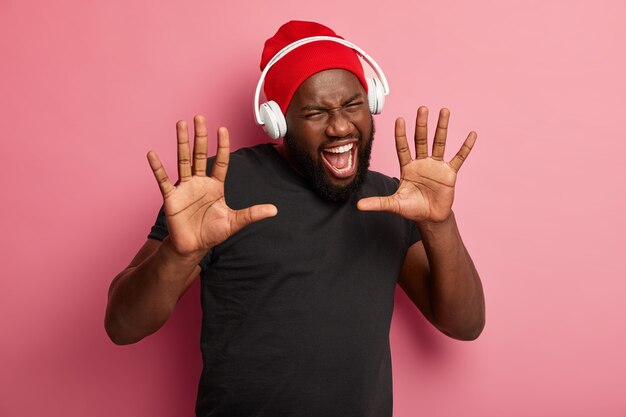 Un homme afro-américain garde les paumes vers l'avant, ouvre largement la bouche, vêtu d'un chapeau rouge et d'un t-shirt noir, s'exclame de joie, aime la musique dans les écouteurs