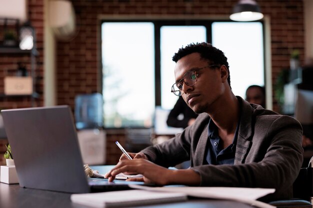 Homme afro-américain handicapé analysant un rapport en ligne sur un ordinateur portable, planifiant une stratégie de projet dans un bureau adapté aux personnes handicapées. Entrepreneur en état de santé assis en fauteuil roulant au travail.