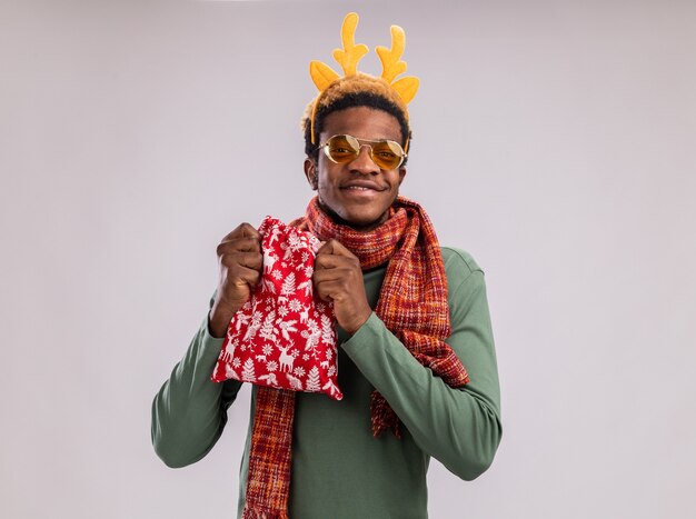 Homme afro-américain avec jante drôle avec des cornes de cerf et une écharpe autour du cou tenant un sac de père Noël rouge avec des cadeaux regardant la caméra heureux et joyeux souriant debout sur fond blanc