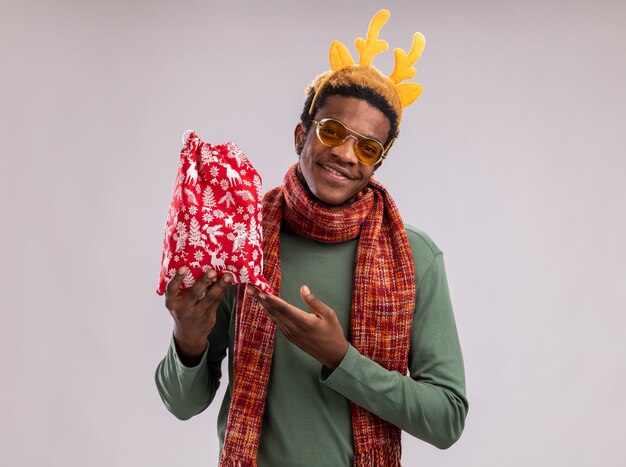 Homme afro-américain avec jante drôle avec des cornes de cerf et une écharpe autour du cou tenant un sac de Père Noël rouge avec des cadeaux regardant la caméra en souriant pointant avec l'index debout sur fond blanc