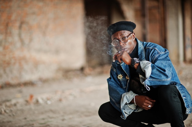Homme afro-américain en jeans veste béret et lunettes fumant un cigare dans une usine abandonnée